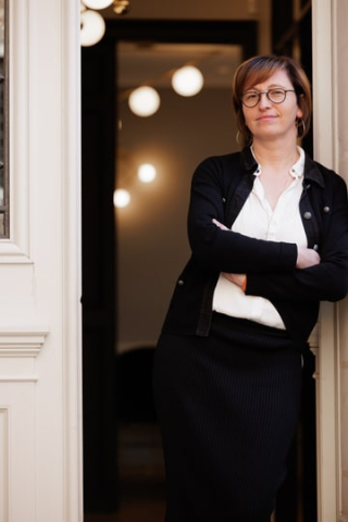Une femme,Aurélie Durand,  debout dans l'encadrement d'une porte, portant un manteau noir et une chemise blanche, les bras croisés, avec un fond de lumières intérieures chaleureuses, dégageant une attitude professionnelle et confiante.pour BLH
