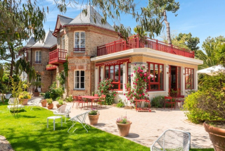 Une maison  en pierre, avec une jolie terrasse, entourée d'arbres verdoyants, sur un fond de ciel bleu clair.