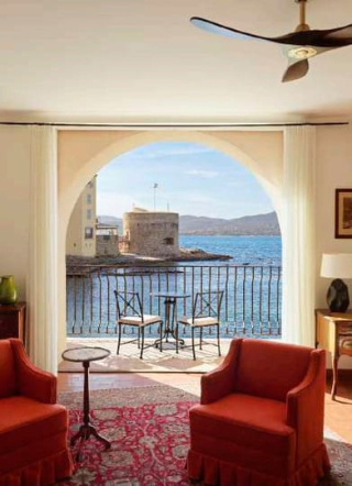 Vue depuis l'intérieur d'une chambre de l'Hôtel La Ponche à travers une arche, donnant sur un balcon aménagé, la mer bleue et la Tour du Portalet de Saint-Tropez au loin.