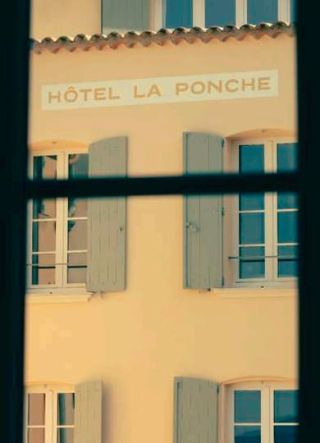 Façade jaune pâle de l'Hôtel La Ponche vue à travers le cadre sombre d'une fenêtre, montrant des volets gris-vert et l'inscription HÔTEL LA PONCHE en lettres capitales.