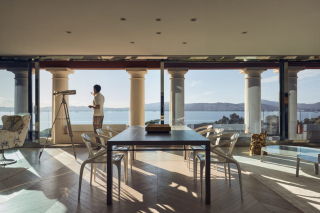 Vue depuis l'intérieur d'une suite luxueuse vers le balcon, où un homme se tient près d'un télescope en regardant la vue imprenable sur la mer.
