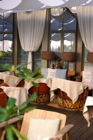 Terrasse de l'hôtel Brach à Paris avec des fauteuils tressés, des plaids blancs et des coussins bleus et blancs portant l'inscription « Beach », sous des voiles d'ombrage blanches.