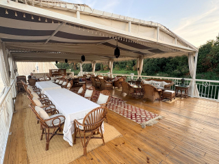 Un salon extérieur élégant et convivial, installé sur une terrasse en bois avec vue sur la verdure, équipé de tables, fauteuils en rotin et banquettes sous une tente ouverte.