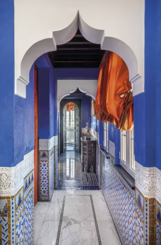 Couloir de La Mamounia aux murs bleu intense, bordé de zelliges, d’arches blanches sculptées et de rideaux orange.
